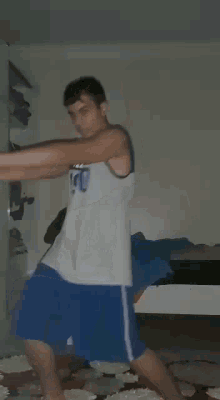 a young man in a white tank top and blue shorts is standing in a bedroom .