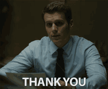 a man in a blue shirt and tie is sitting at a desk with a thank you sign behind him