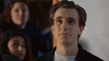 a man in a black jacket looks at the camera with a woman in the background