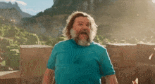 a man with a beard and long hair is wearing a blue shirt and standing in front of a mountain .