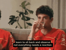 a man in a red shirt is sitting on a couch drinking a glass of water and saying " learn to sit back and observe "