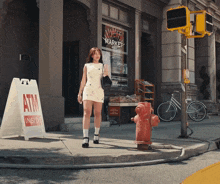 a woman in a white dress walks down a sidewalk in front of an atm sign