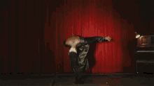 a man in a grey dress is standing in front of a red curtain with his arms outstretched