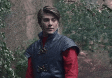 a man in a leather vest and red shirt stands in front of trees