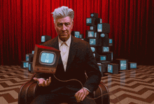 a man in a suit is sitting in front of a stack of televisions and holding a small television