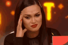 a woman with long black hair and red lipstick is sitting at a table with her hand on her forehead .