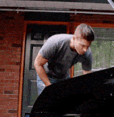 a man in a gray shirt is looking under the hood of a car