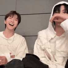 two men are sitting next to each other and laughing . one of the men is wearing a hooded jacket .