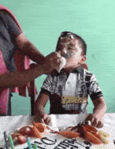 a little boy is sitting at a table with a birthday cake and a person is icing his face .