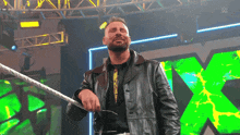 a man in a leather jacket is standing in a wrestling ring with a green x in the background