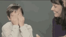 a young boy covering his eyes with his hands while a woman smiles behind him