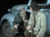 two men are sitting next to a blue car