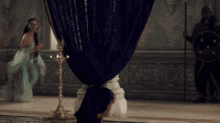 a woman in a blue dress stands in front of a blue curtain while a man with a shield stands behind her