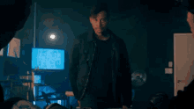 a man in a black jacket is standing in a dark room with a computer monitor in the background .
