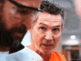 a man with a beard wearing glasses looks at another man in an orange uniform