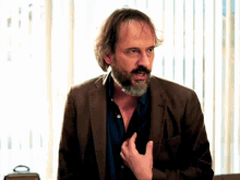 a man with a beard wearing a brown jacket is standing in front of a window