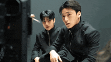 a man in a black jacket sits next to another man