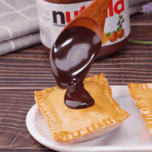 a bottle of nutella is behind a wooden spoon pouring chocolate sauce on a pastry
