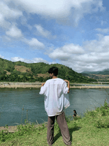 a man in a white shirt stands on a grassy hill overlooking a river
