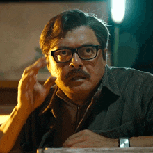a man with glasses and a mustache is sitting at a table with a watch on his wrist