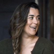 a woman wearing a green sweater and a necklace is smiling with her eyes closed