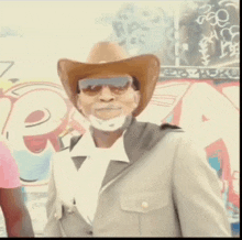 a man wearing a cowboy hat and sunglasses stands in front of a graffiti wall with the letter e on it
