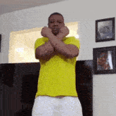 a man in a yellow shirt and white shorts is standing in a living room with his hands crossed .