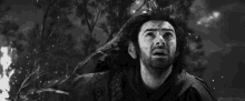 a man with long hair and a beard is standing in front of a tree looking up at the sky .