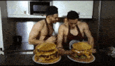 two shirtless men are standing next to each other in a kitchen eating huge hamburgers .