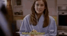 a woman in a robe is saying merry christmas while standing in a kitchen .