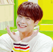 a young man wearing a white striped sweater smiles with his hands on his face