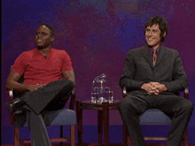 a man in a suit sits next to another man in a red shirt on a stage