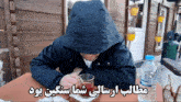 a man in a hooded jacket sits at a table with a bottle of water and a glass of tea