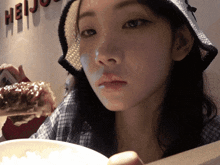 a woman wearing a hat is eating a piece of food in front of a wall that says heiju