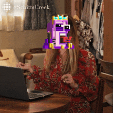 a woman is sitting at a table with a laptop and a purple pixelated face behind her
