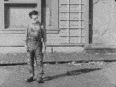a black and white photo of a man standing on the sidewalk in front of a building .