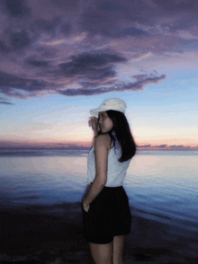a woman wearing a white hat stands in front of the ocean at sunset