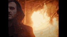 a man in a hooded jacket stands in front of a burning building