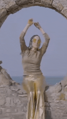 a woman in gold pants and sunglasses is standing in front of a stone archway overlooking the ocean .