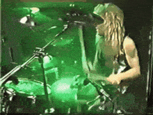 a man with long blonde hair is playing drums in front of a green light