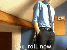 a man in a blue shirt and tie is standing in front of a wall that says you roll now