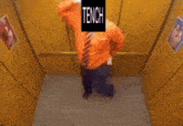 a man in an orange shirt and tie is standing in an elevator with a sign that says tench above him