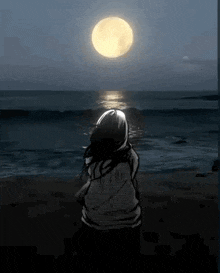 a woman is standing on a beach looking at the full moon .
