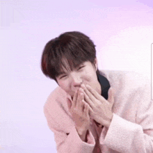 a young man in a pink sweater is making a funny face with his hands covering his mouth .