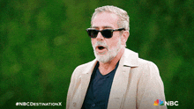 a man with a beard wearing sunglasses and a trench coat with the nbc destination x logo