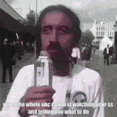 a man with a mustache is holding a bottle of water and talking to someone .