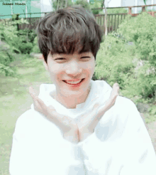 a young man wearing a white hoodie is smiling and making a heart with his hands .