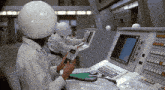 a man wearing a white helmet sits at a control panel holding a phone