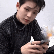 a young man wearing a black sweater and a silver necklace is looking at his cell phone .