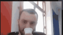 a man with a beard is drinking from a cup in front of a window